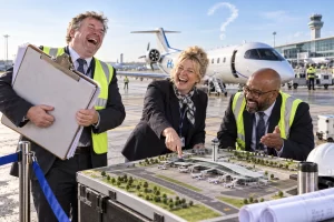 Three fictional adult airport officials clearly laugh beside a hydrogen aircraft and an airport expansion model on a runway.