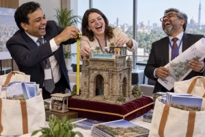 Three laughing conference delegates measure a model stone gateway topped with a miniature pool beside piles of tourism brochures.