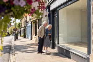 Original editorial photograph of two laughing older adults beside a plain empty shop window on a sunny British high street, with no signs or text