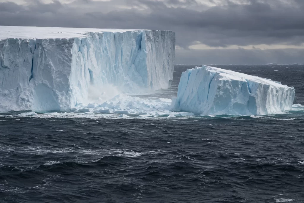 A newly separated slab of pale blue ice floats beside a colossal Antarctic iceberg in dark choppy ocean water.