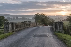 A weathered rural bridge carries an empty road past a shallow repaired section and plain safety cones.