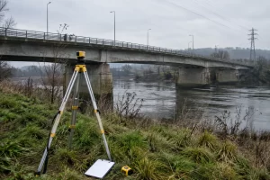 A survey tripod and blank clipboard sit beside a concrete bridge crossing a river under an overcast sky.