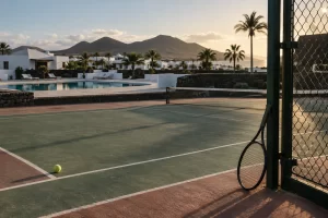 An empty resort tennis court beside a still pool has a lone racket and ball in late island sunlight.