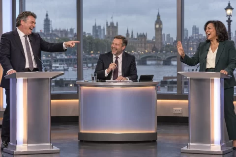 Satirical illustration of two laughing politicians debating in Westminster with Big Ben visible through a rainy window.