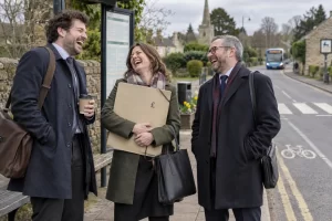 Three fictional adult commuters clearly laugh at a rural bus stop while holding a large funding folder and waiting for a bus in the distance.