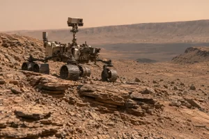 A six wheeled robotic Mars rover travels over layered reddish rock ridges beneath a pale salmon sky.