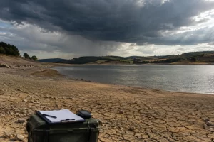 A low reservoir with cracked shoreline sits below a dense rain cloud over the English countryside.