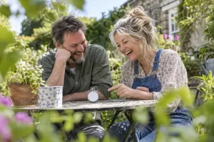 Original editorial photograph of two laughing adults beside a small blank stopwatch in a sunny community health garden for motor neurone disease fast tracked care