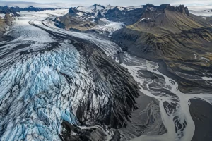 Pale blue glacier crevasses, dark volcanic ash streaks and a winding glacial river cross an Icelandic highland valley.