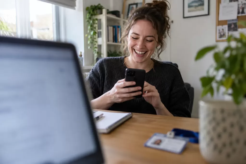 One laughing adult new employee looks at a generic smartphone in a home office, framed through a blurred laptop screen.