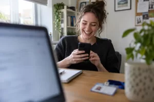 One laughing adult new employee looks at a generic smartphone in a home office, framed through a blurred laptop screen.
