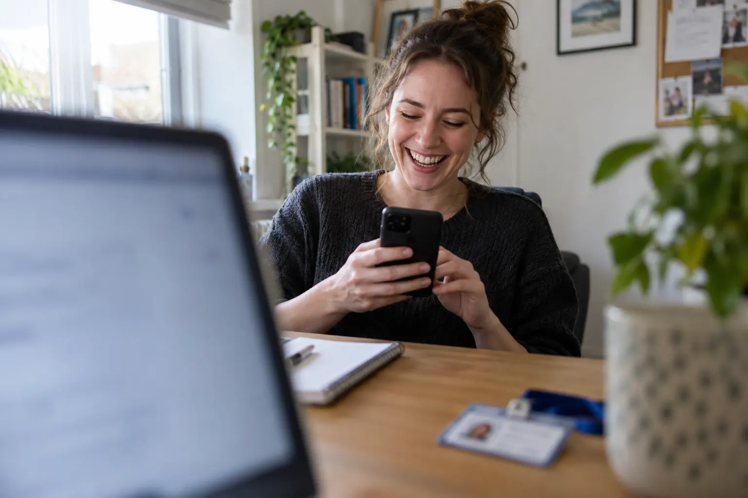One laughing adult new employee looks at a generic smartphone in a home office, framed through a blurred laptop screen.