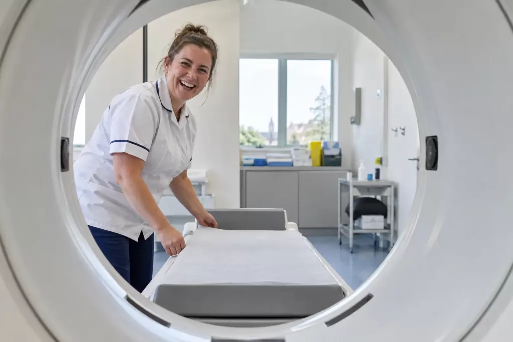 One laughing adult radiographer appears through the circular opening of a generic bone scanner in an empty scan room.