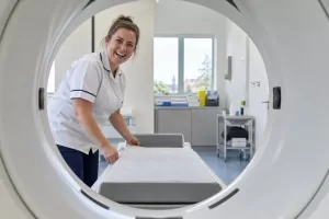 One laughing adult radiographer appears through the circular opening of a generic bone scanner in an empty scan room.