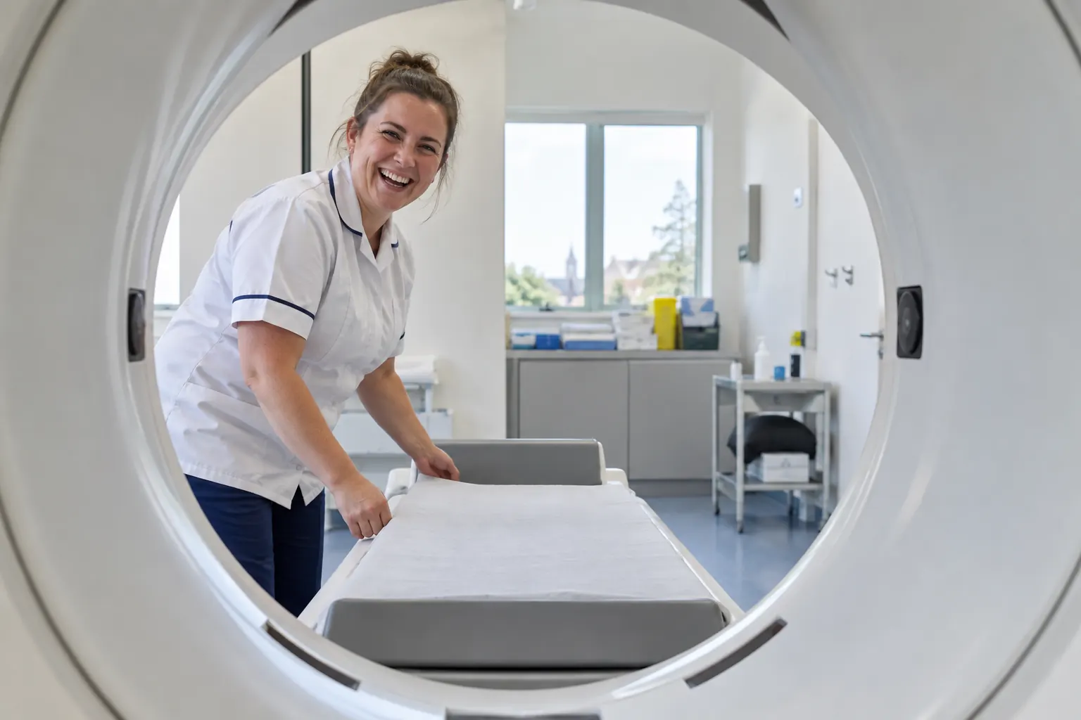One laughing adult radiographer appears through the circular opening of a generic bone scanner in an empty scan room.