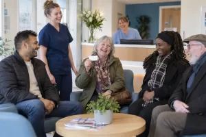 Six laughing adult patients and dental staff in a bright NHS-style dental waiting room.