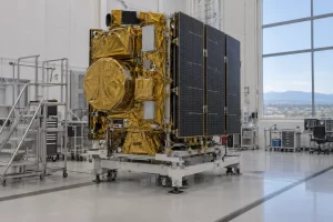 A gold wrapped Earth observation satellite with solar panels stands in a bright aerospace clean room.