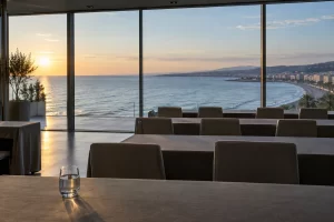An empty waterfront conference room looks out over calm Mediterranean water at sunrise.