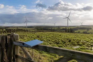 Wind turbines turn across green farmland behind a wooden gate holding a plain blue planning folder.