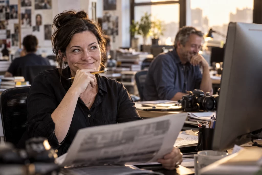 Newspaper editor smiling wryly while reviewing an unreadable printed page in a newsroom