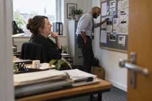 Two laughing adult public-sector fraud investigators work at different depths in an office, viewed from behind an open door.