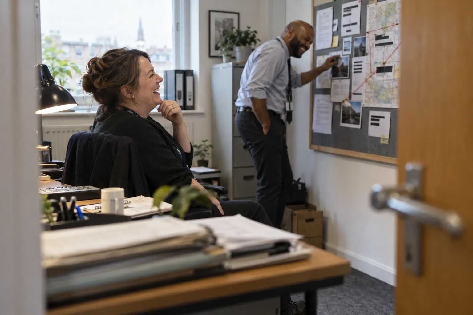 Two laughing adult public-sector fraud investigators work at different depths in an office, viewed from behind an open door.