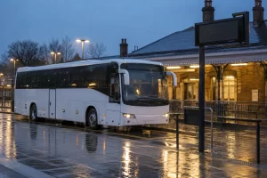 An unbranded coach waits beside a blank information board outside a rain wet railway station at dusk.