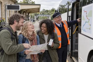 Three laughing adult passengers and a laughing adult dispatcher study a route display beside a rail replacement coach at a fictional railway station.