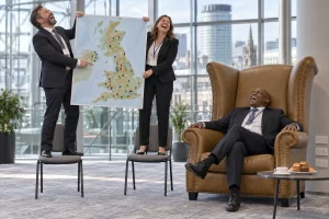 Two laughing delegates stand on small chairs holding a map while a third laughs from an oversized chair beside coffee and power sockets.