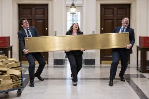 Three laughing officials carry an enormous blank brass nameplate between identical office doors beside a trolley of smaller plates.