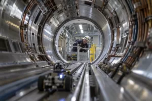 One laughing adult engineer peers into a vast accelerator tunnel while a generic mouse-sized inspection robot sits close to the camera.