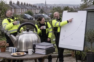 Three fictional police officers laugh around an oversized kettle and blank tactical chart in a suburban garden.
