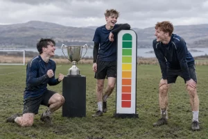 Three laughing young football players stand beside a plain trophy and a blank coloured optimism meter on a muddy pitch.