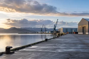 An empty Scottish industrial harbour has calm water cranes warehouses and distant hills at first light.