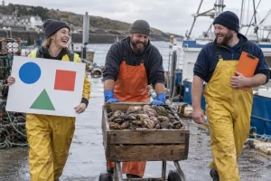 Three laughing seafood export workers wheel a crate of shellfish to a cargo ship beside a blank presentation board.