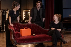 Three laughing theatre staff stand beside a velvet fainting sofa with a plain orange emergency kit and bell.
