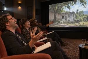 Three laughing festival programmers watch a cinema screen showing a quiet tropical landscape with a wall, goat and parked motorbike.