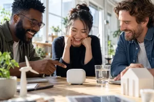 Original editorial photograph of three laughing innovators around a plain smart meter and glass of water in a sunny studio, with no logos or text
