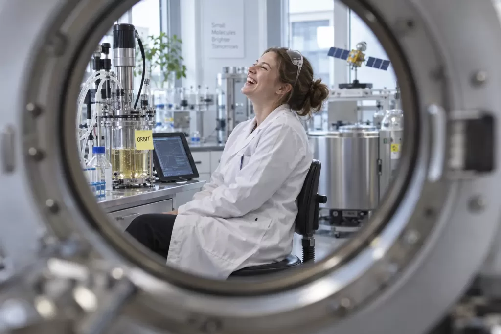 A laughing pharmaceutical scientist sits beside a laboratory bioreactor, seen through the circular opening of a large scientific instrument.
