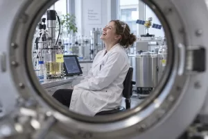 A laughing pharmaceutical scientist sits beside a laboratory bioreactor, seen through the circular opening of a large scientific instrument.