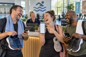 Three fictional adults clearly laugh at a leisure centre reception while one holds a smartphone beside their trainers and a receptionist laughs behind the desk.