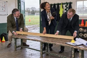 Three fictional sports funding officials laugh while measuring a battered changing room bench.