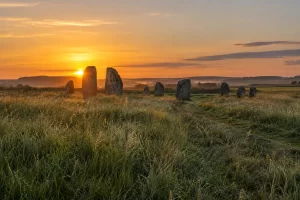 Sunrise shines through a distant ancient stone circle above a dewy English grassland on the longest day.