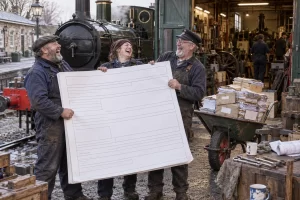 Three fictional adult railway volunteers laugh beside a heritage steam locomotive while an oversized grant application blocks a workshop entrance.