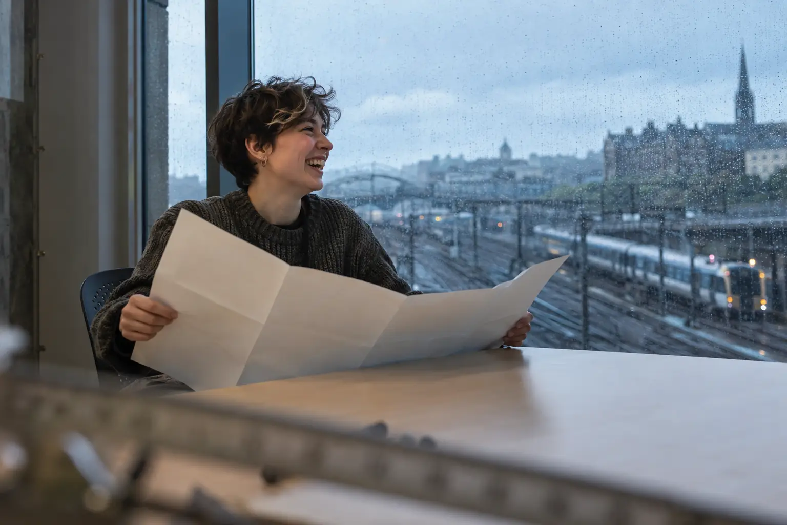 Original editorial photograph of a laughing young non-binary transport modeller alone at a rainy window, holding a blank paper map with no text