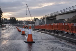 A long line of orange traffic cones and temporary barriers leads past wet roadworks and a new rail bridge at dawn.