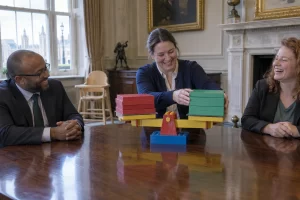 Three laughing civil servants balance blank coloured folders on a toy block scale in a formal meeting room.