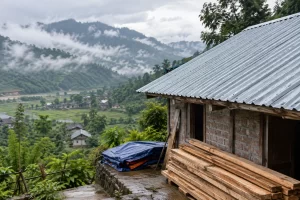 A repaired metal roof and stacked timber stand beside a brick house in a misty mountain valley.