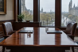 Two empty leather chairs face one another across a meeting table with plain folders and water glasses on a rainy day.