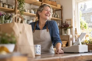 Original editorial photograph of a laughing middle-aged woman shopkeeper alone beside a blank small cash register in a sunlit shop with no text
